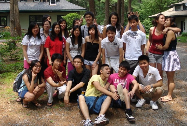 A group of 21 teens or young adults at an outdoor retreat in a forest with a few house-like buildings behind them. They are having fun posing the for photo, some giving their peers bunny ears or making funny faces.