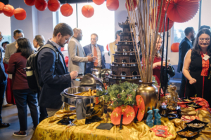 Attendees at the Lantern Festival Gala select appetizers from a table decorated with various festive Chinese items, like red fans, bamboo in a gold vase, and guardian lion statues.