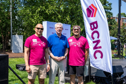 At the outdoor Family Fun Fair in Quincy, 2 volunteers and a sponsor pose happily for a photo together by a flag with BCNC's logo.